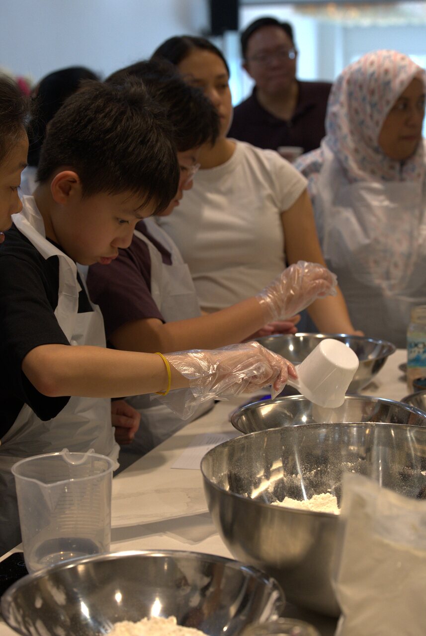 A young participant measuring flour into a mixing bowl with help from the people beside him
