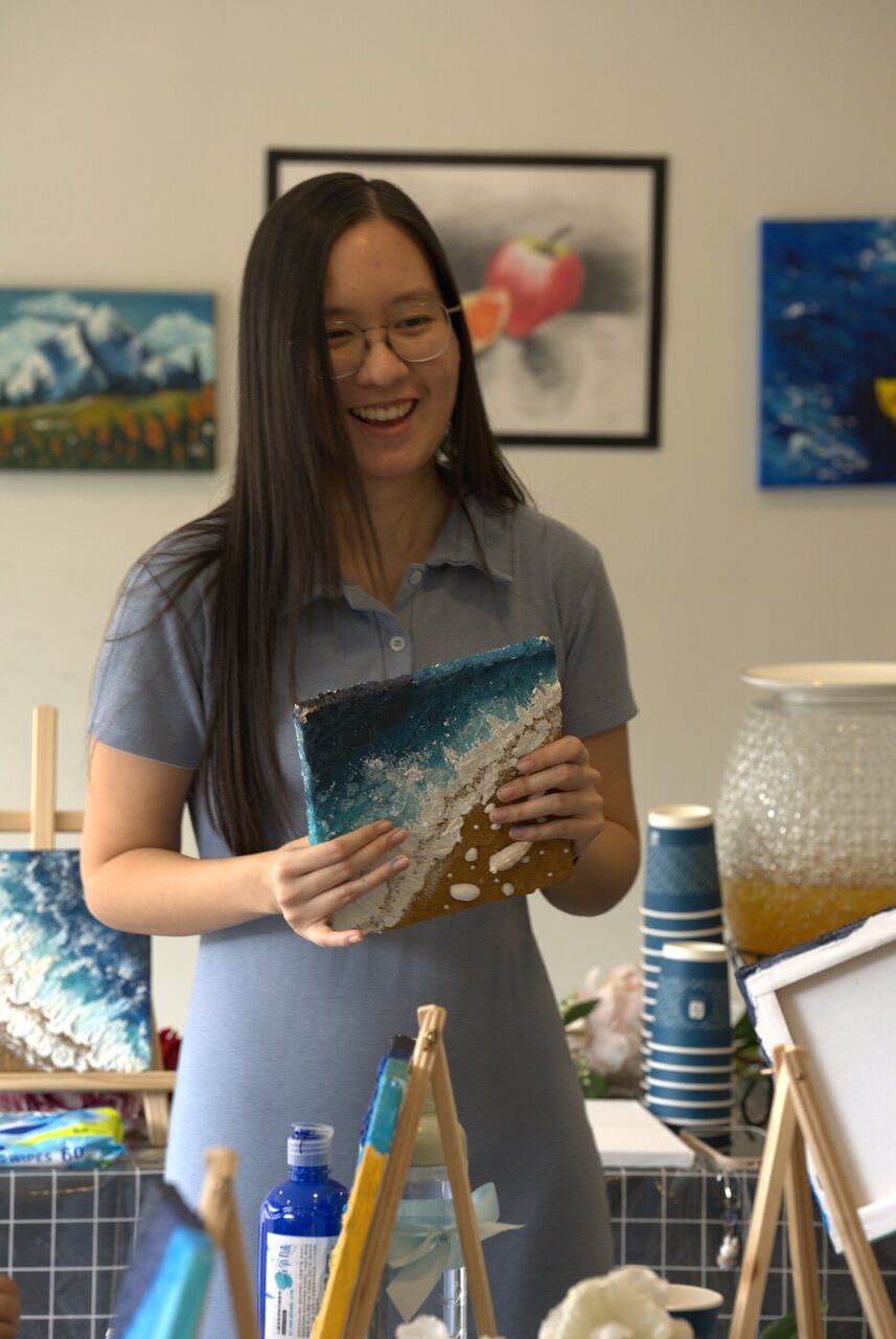 A painter smiling while holding up her finished textured shoreline canvas